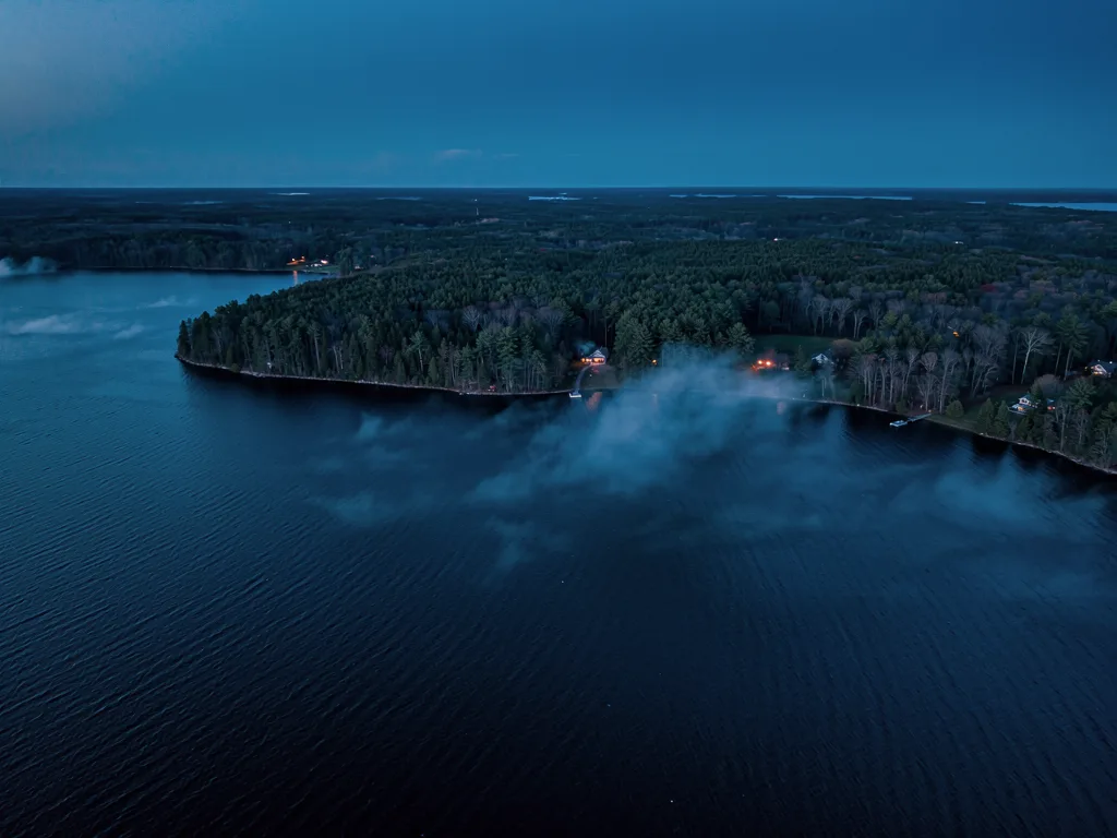 Aerial view at dusk of a forested point on a large lake, a few lit windows among the pines along the shoreline and low mist drifting over dark water