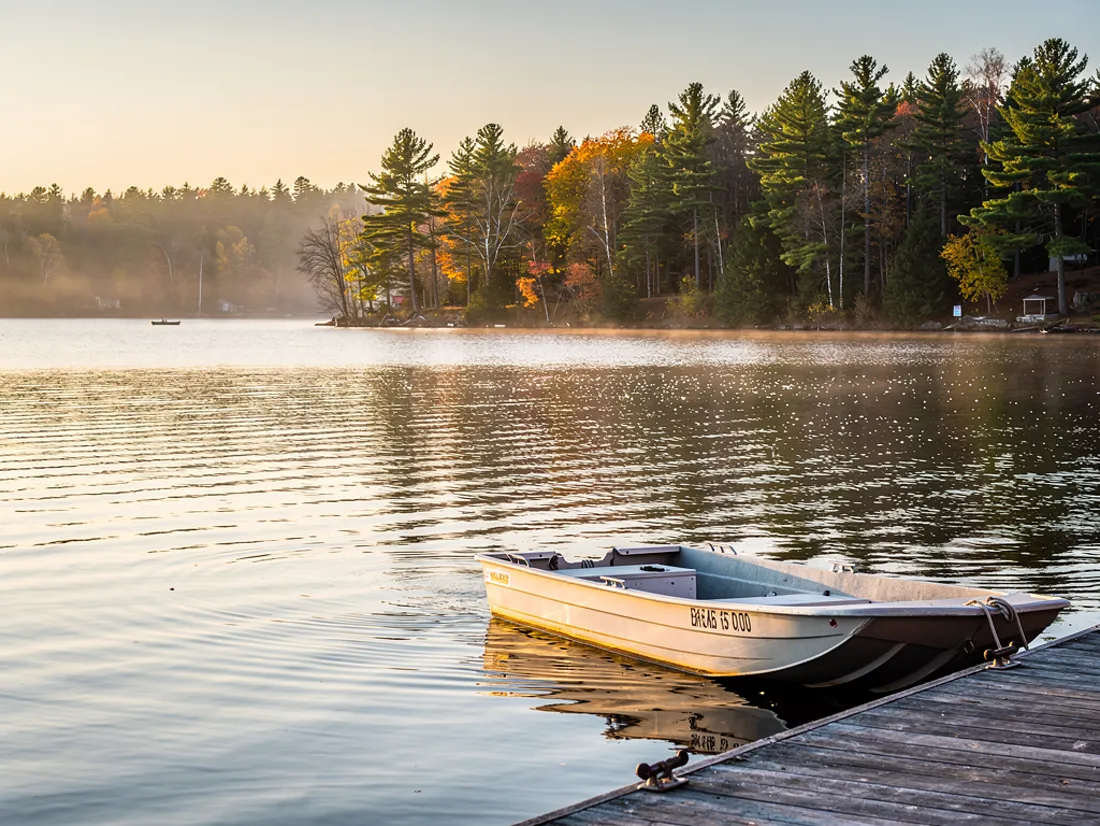 An aluminum fishing boat tied to a wooden dock on a calm Northern Michigan lake, autumn color on the far shore