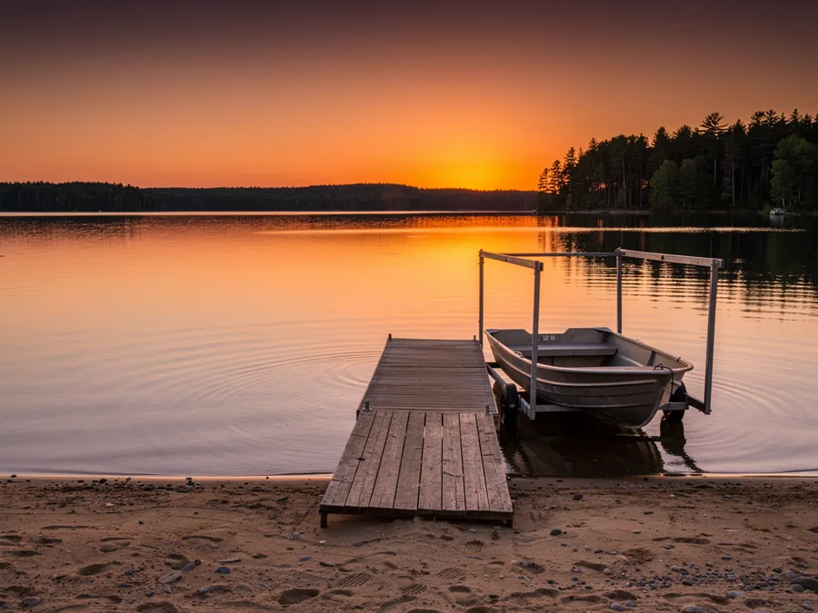 A wide inland lake at sunset, a wooden dock and boat hoist on a sandy shoreline with pine forest beyond